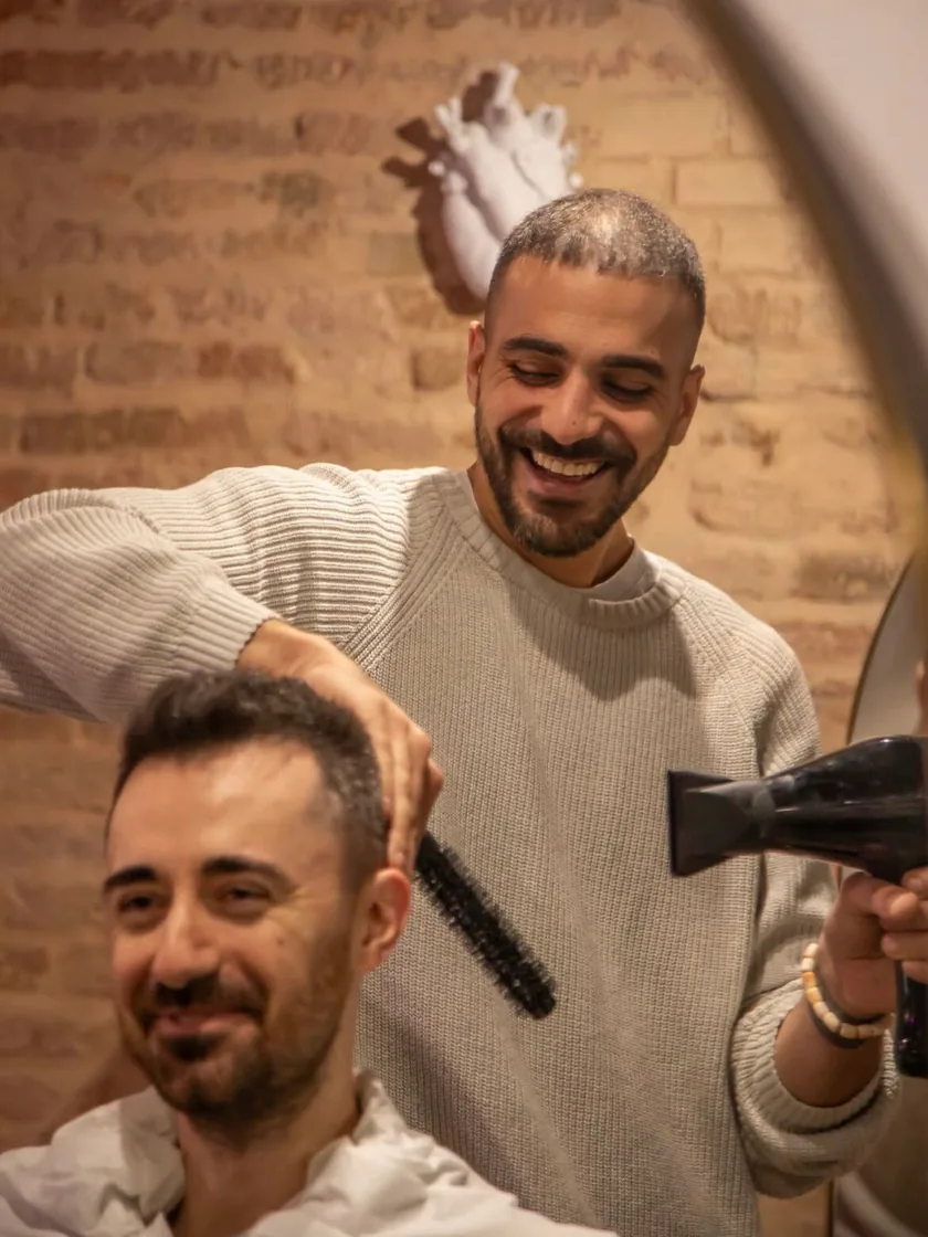 Enrico Cantore ride mentre asciuga i capelli a un cliente con phon e spazzola tonda, nel salone di Via Saragozza